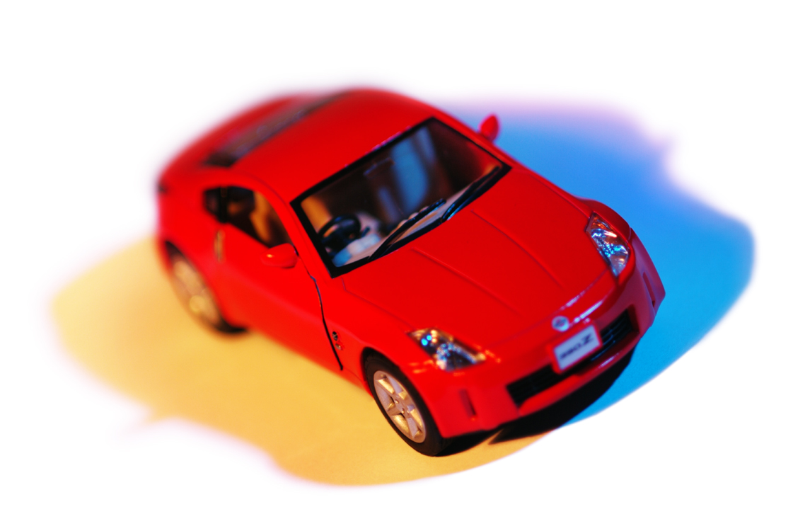 A red toy car model resembling a sports car. It's photographed with a colorful light effect, casting shadows with hints of blue and yellow on the surface. The model features detailed headlights and a visible windshield, adding to the realistic look of the miniature vehicle.